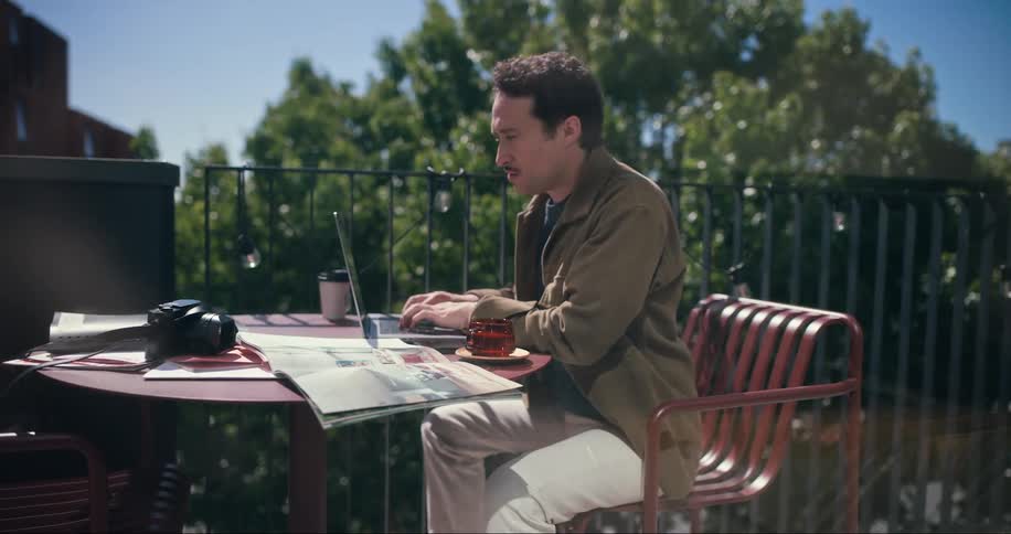 Man sits at an outdoor table typing on a laptop after consulting a publication