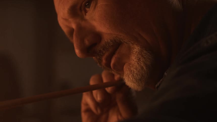 A man with a gray beard is holding a twig in his hand looking straight ahead concentrating