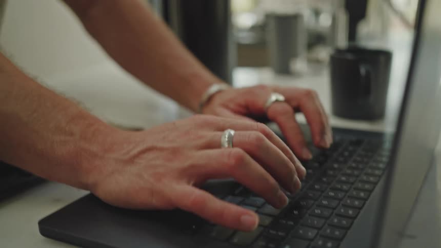 A blonde man hands with rings and a bracelet typing on a laptop keyboard with a partially visible mug in the background