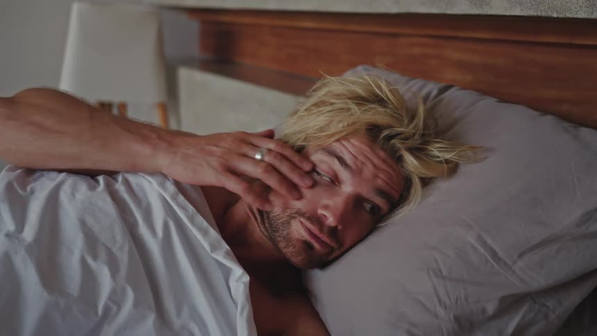A blonde shirtless man lying in bed wakes up and sits up in the bed in the bedroom