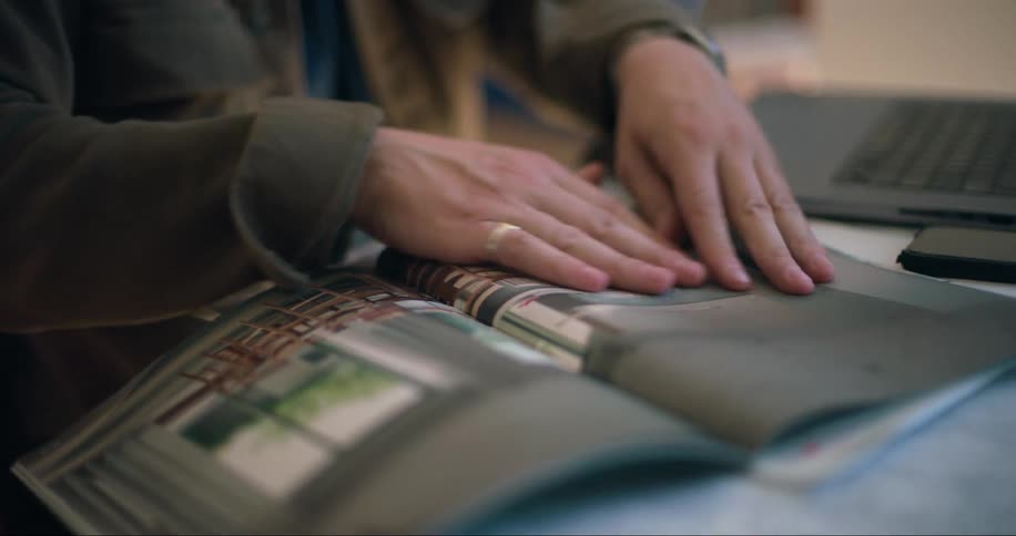 A person working on a laptop at home flips through the pages of a magazine beside it