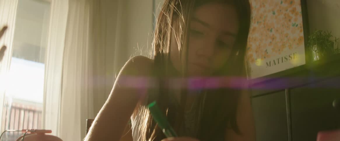 A girl with long hair is sitting at a table drawing with colored pencils in a sunlit room with a window