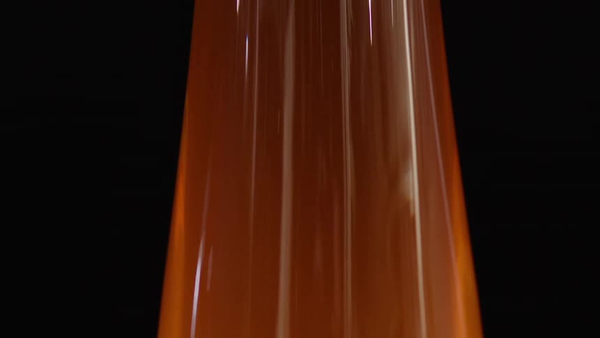 A viscous transparent blob of molten glass is being stretched vertically against a dark background