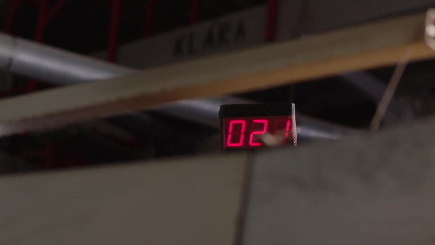 A digital display with red numbers is mounted on a structure with industrial pipes in the background