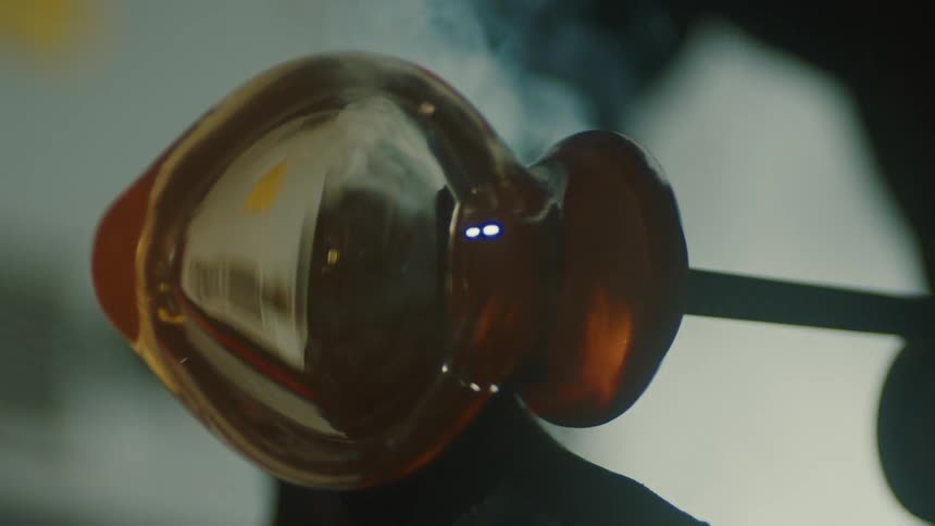 A piece of molten glass is being shaped and cooled on a surface with smoke visible in the background capturing the glassblowing process