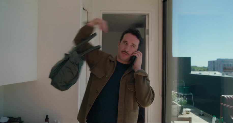 A man on the phone picks up a handbag while walking through an apartment seemingly ready to go out