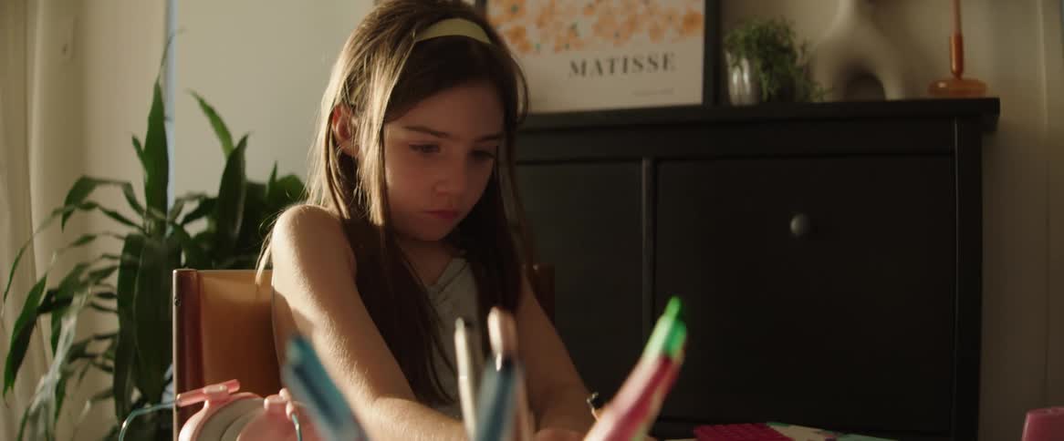 A girl with long hair is sitting at a table drawing with colored pencils in a sunlit room