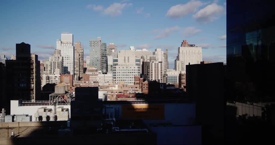 A cityscape skyline stretches across the frame showcasing a variety of buildings under a bright blue sky dotted with fluffy white clouds