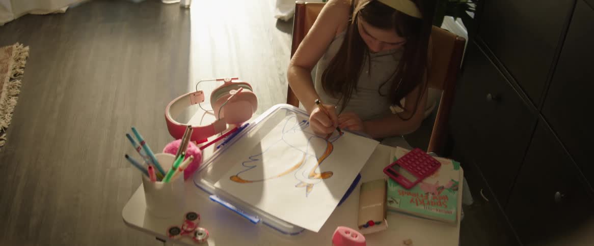 A girl with long hair is sitting at a table drawing with colored pencils in a sunlit room with a window