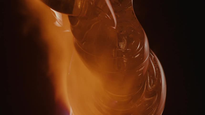 Viscous golden material being heated with a torch likely in a glassblowing or similar process against a dark background