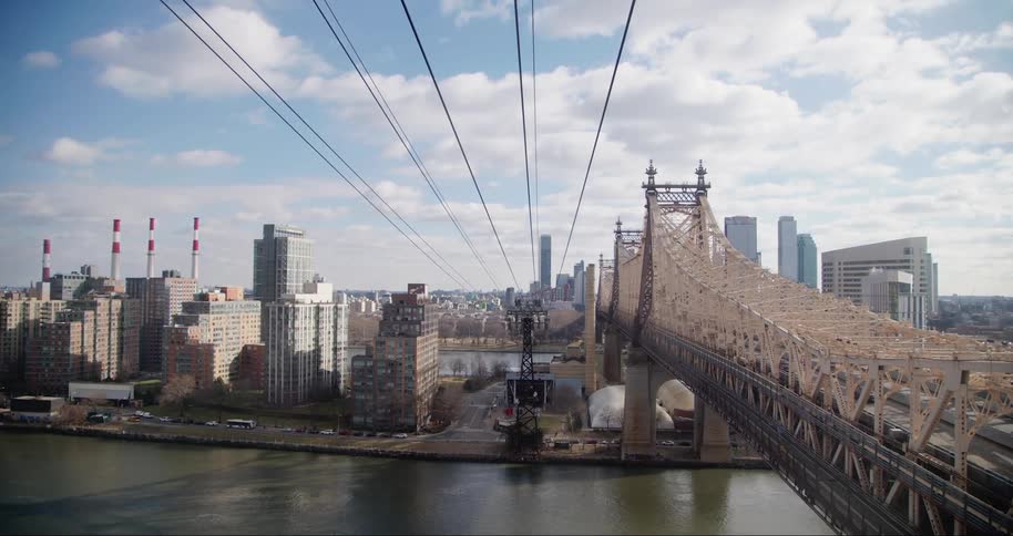The roosevelt island bridge spans the east river toward the cityscape of manhattan and the ravenswood generating station smokestacks as seen from a cable car