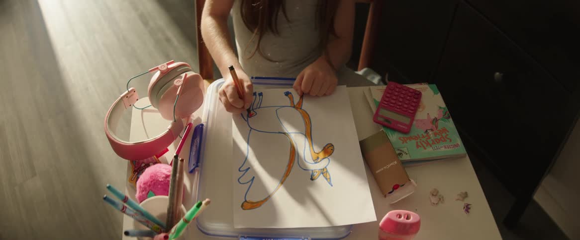 A young girl sits at a table drawing a colorful whimsical creature surrounded by art supplies and personal items