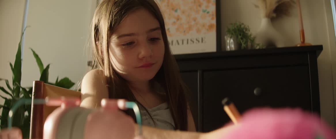 A girl with long hair is sitting at a table drawing with colored pencils in a sunlit room