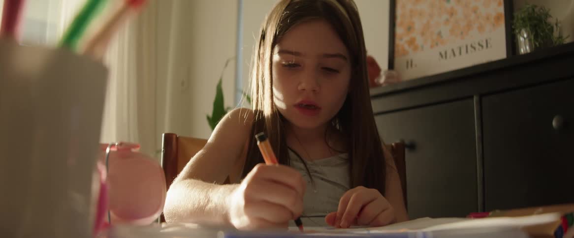 A girl with long hair is sitting at a table drawing with colored pencils in a sunlit room with a window