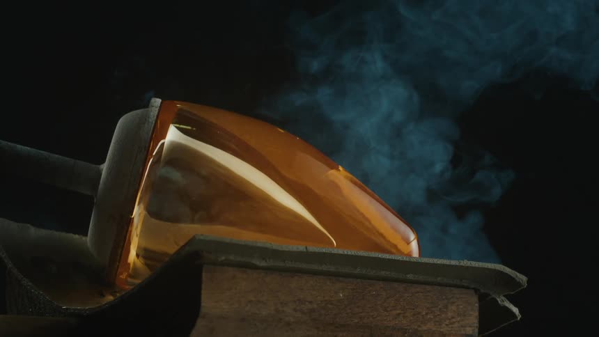A piece of molten glass is being shaped and cooled on a wooden surface with smoke visible in the background capturing the glassblowing process