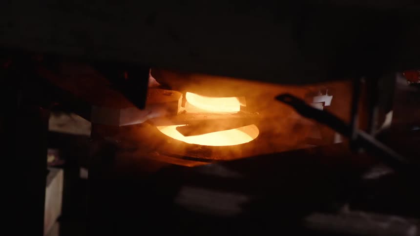 Closeup of an industrial machine with a stream of molten glass pouring into a metallic container being cut by a pair of metal tools
