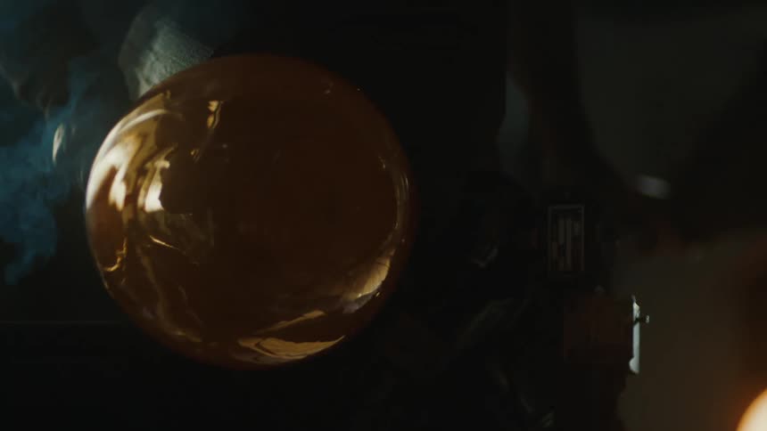 A piece of molten glass is being shaped and cooled on a wooden surface with smoke visible in the background capturing the glassblowing process