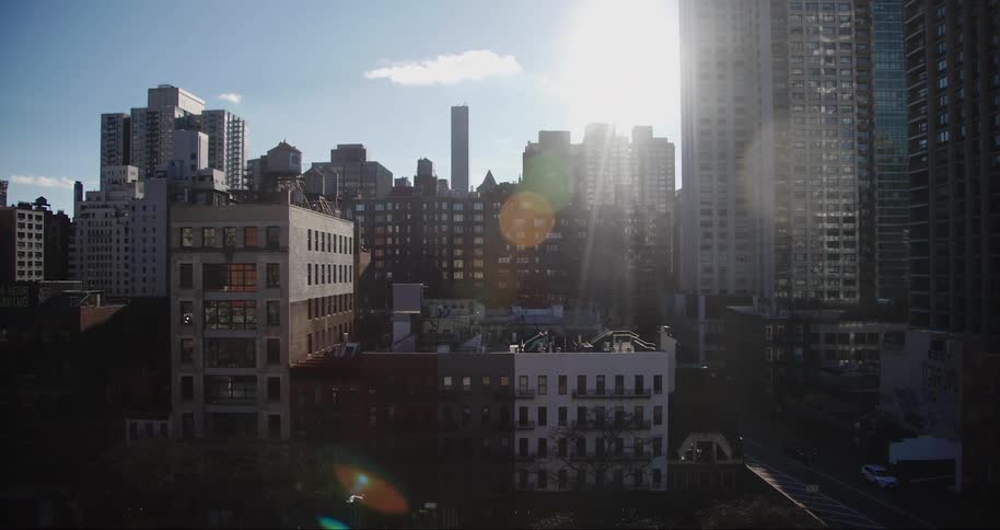 The sun shines brightly over a cityscape illuminating a variety of buildings ranging from shorter older structures to modern high rises with the sunlight creating a bright spot in the sky and casting long shadows across the urban scene