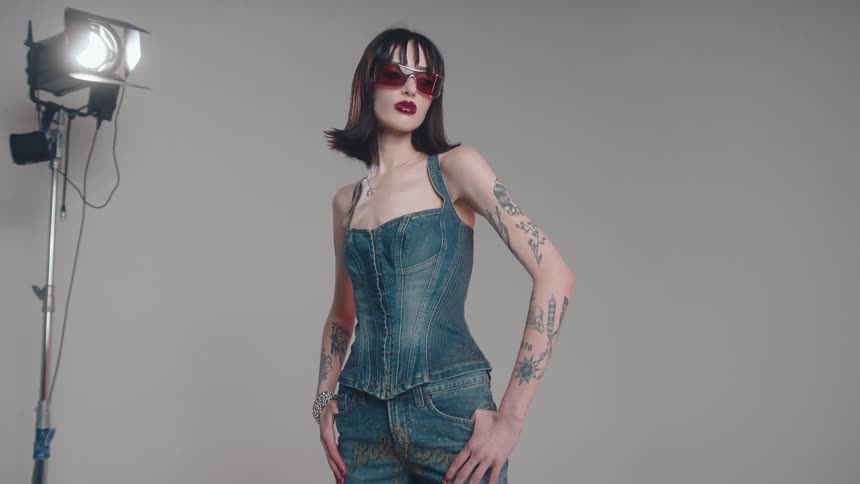 A tattooed model in denim poses in a studio with a lighting setup