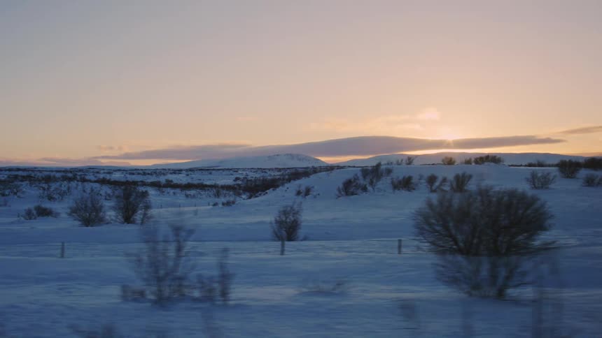 Snowy landscape with sparse vegetation and snowcapped hills at sunrise with the sun on the horizon