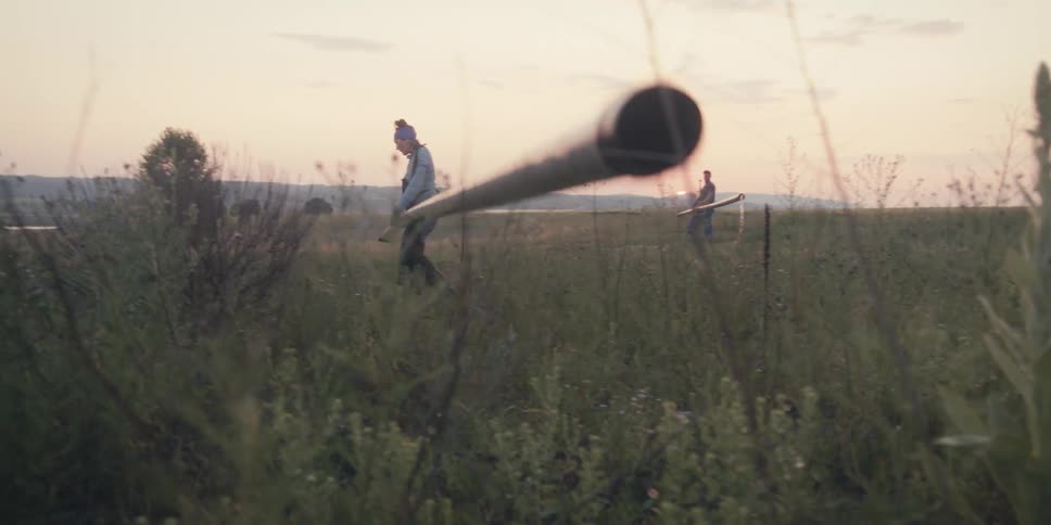 Two people carry a long irrigation water pipe across a grassy field at sunset surrounded by wild plants and soft warm light as they walk down the hill