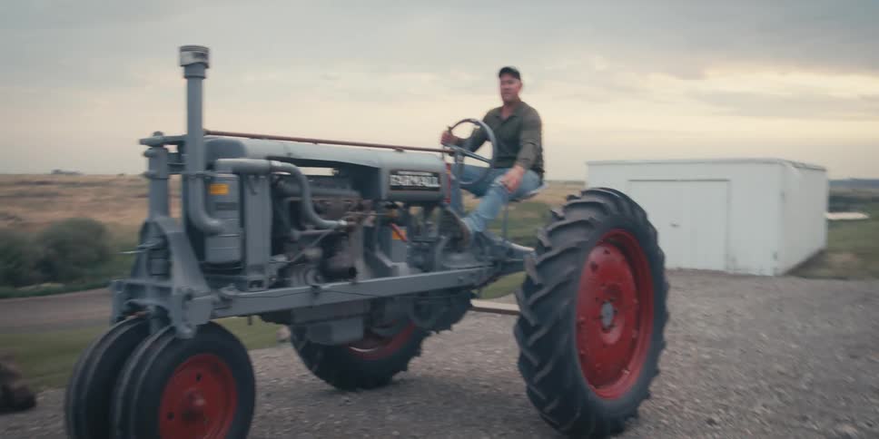 A person is sitting on a vintage farmall tractor with large red wheels parked on a gravel path with a white building and expansive fields in the background