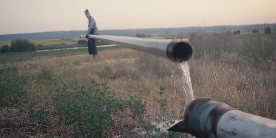 A woman is trying to attach a long pipe to a water source on a farm
