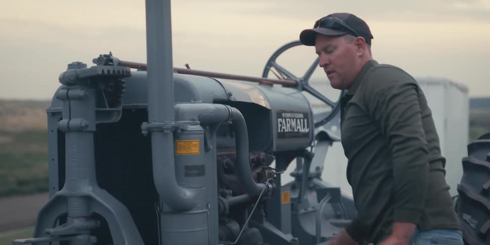 A farmer attempts to handcrank start a vintage tractor in a rural field during the evening with rolling farmland and soft light in the background