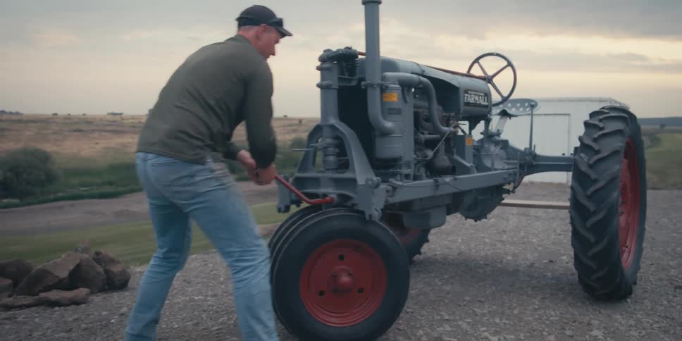 A man is shown handcranking a vintage farmall tractor on a rural farm on a cloudy overcast day