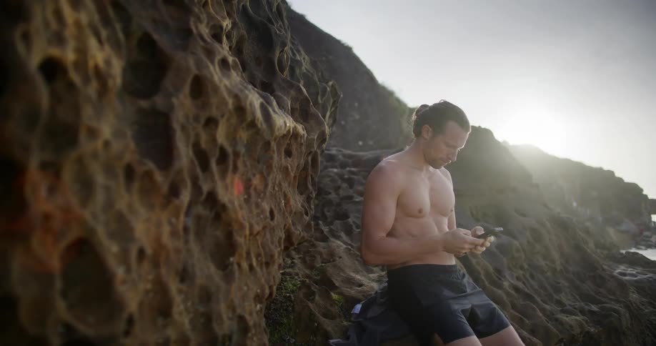 Man in shorts and no shirt sitting on rocky stones by the sea and typing on a cell phone