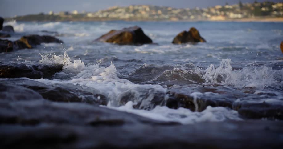 Waves crash against dark rocks in the foreground with a blurred coastline and city skyline in the distance