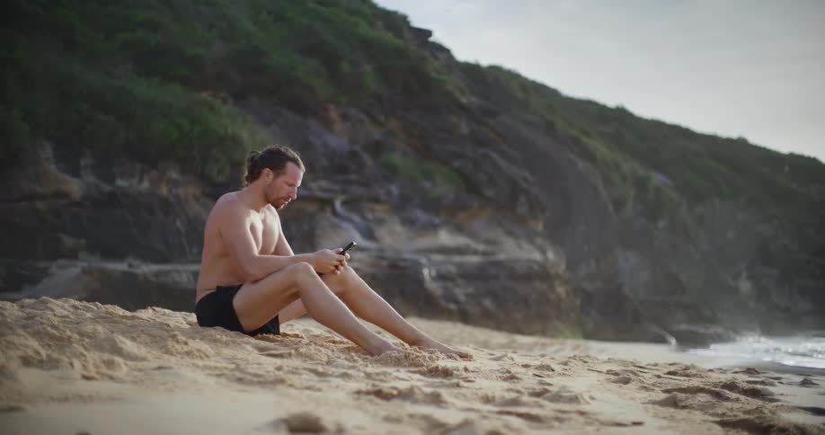 Man sitting on the sand at the beach using his cell phone