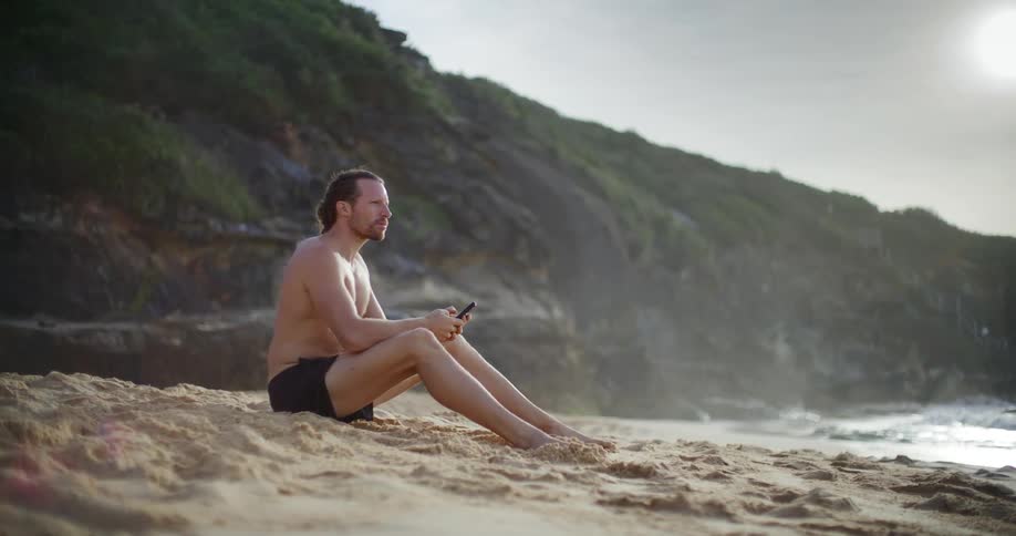 Man sitting on the sand at the beach interacting with a smartphone