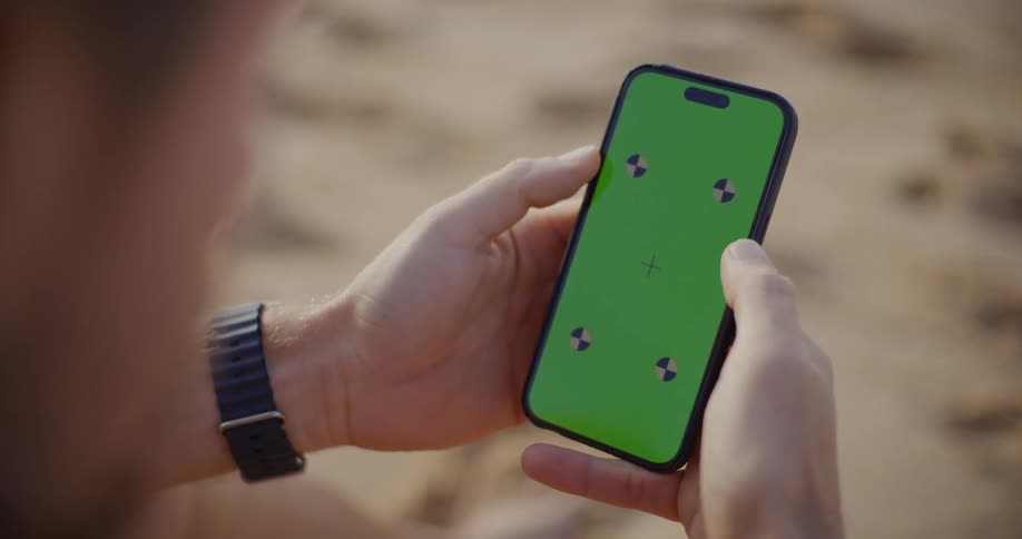 A persons hands are holding a smartphone with a bright green screen displaying four small black tracking markers for chroma key compositing with a blurred background