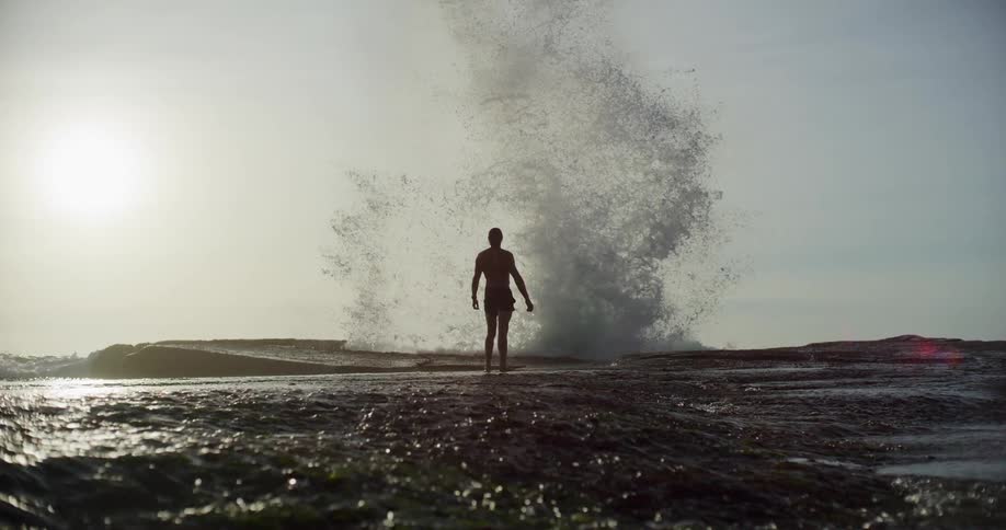 Man standing with his back turned in a natural area with waves crashing into rocks and splashing upwards