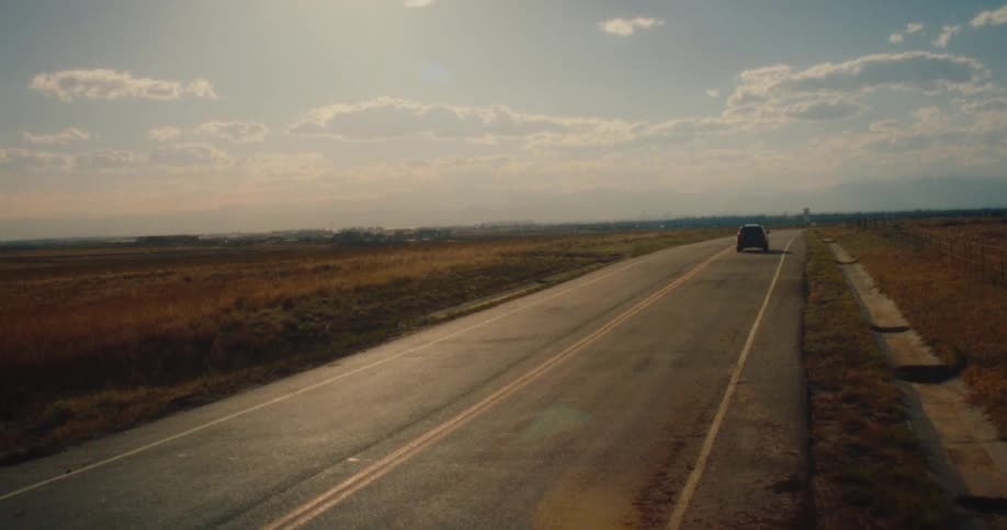 A lone vehicle drives down a straight road that cuts through a vast dry landscape under a partly cloudy sky