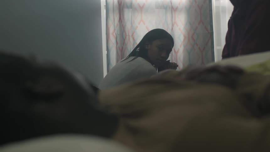 A woman sits in thought in a bedroom where a man is lying on a bed
