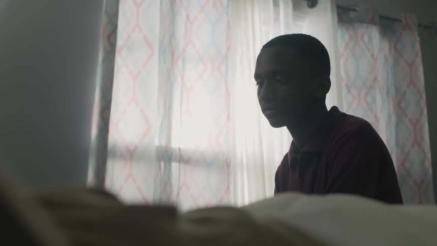 A boy sits near the window nodding to someone in bed