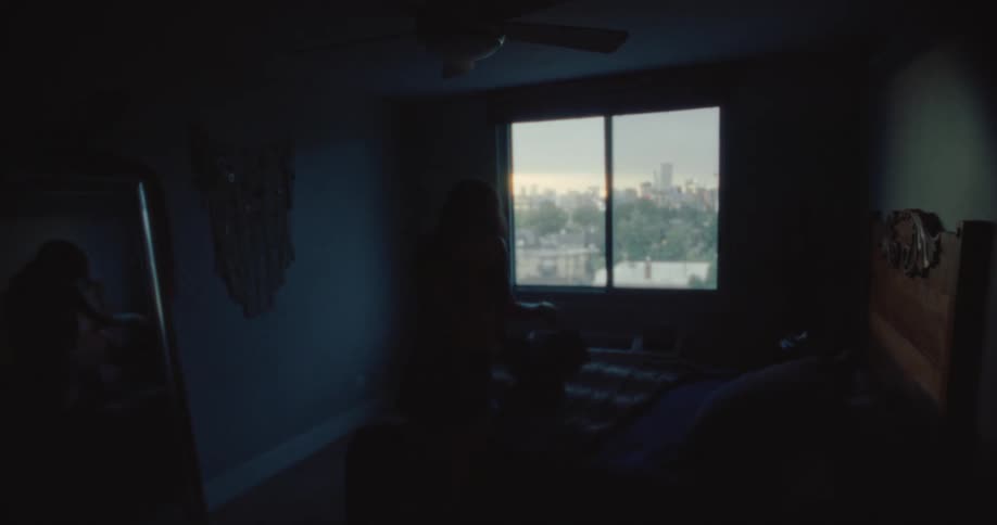 A woman packing clothes on a bed in a dimly lit room with a window overlooking the city