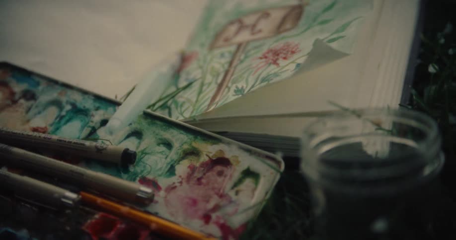 A partially filled watercolor palette with brushes rests beside an open sketchbook displaying a painted floral design with a glass of water nearby