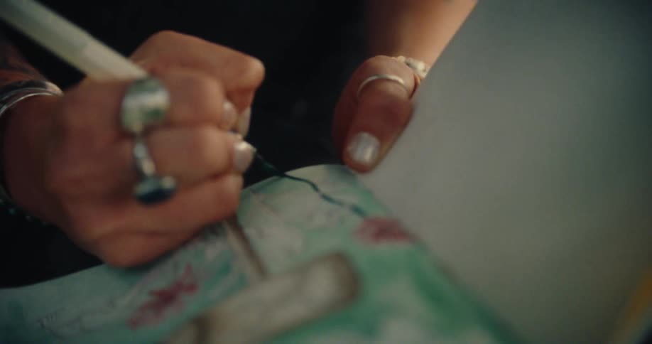 A hand adorned with rings and bracelets delicately paints a detailed design on a piece of paper