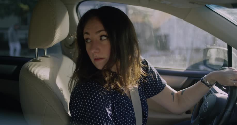 A smiling woman in a car puts on her seatbelt then reverses and drives away down the street