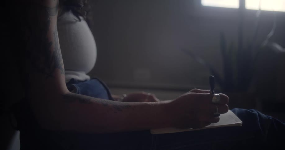 A woman with a tattooed arm sits in a dimly lit room writing in a notebook