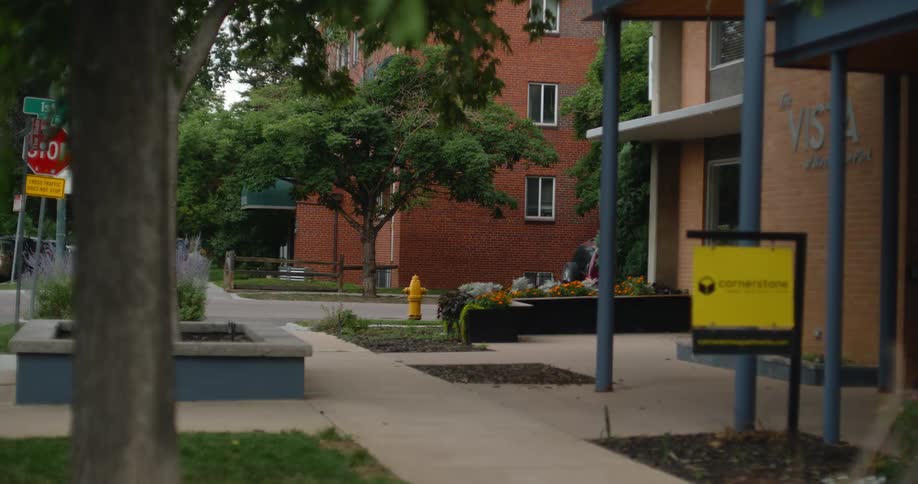 A multistory brick building with whitepaned windows stands behind a paved walkway dotted with trees flowerbeds and urban signage