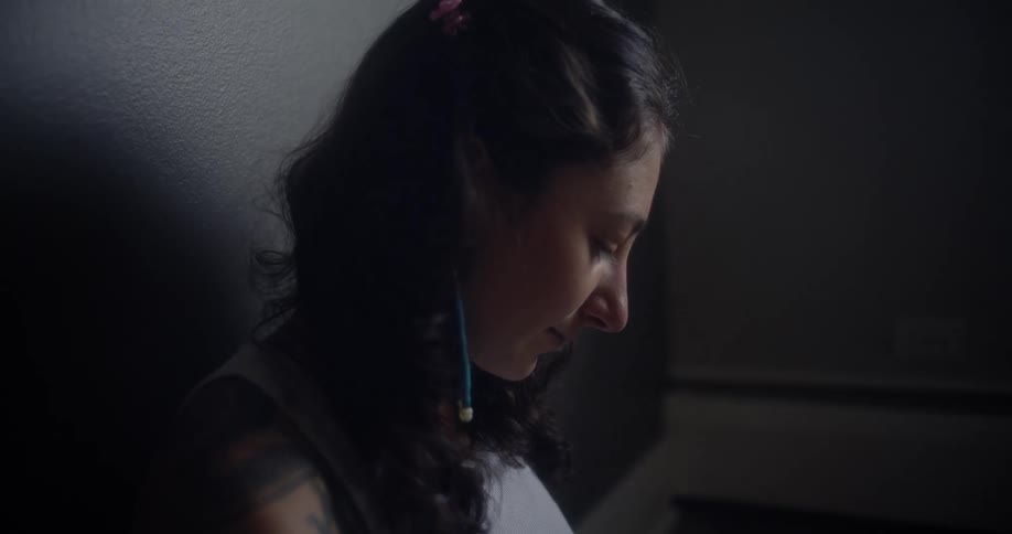 A woman with a tattooed arm sits on the floor in a dimly lit room writing in a notebook