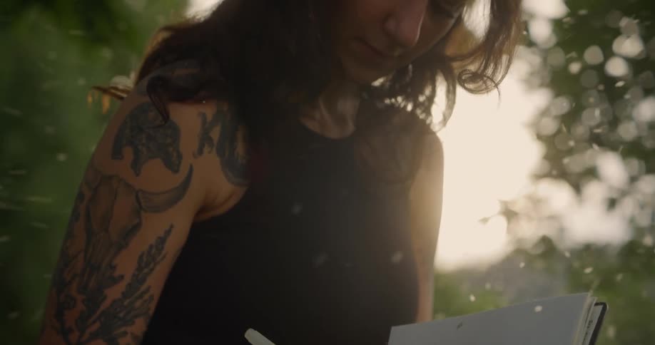 A woman with long hair and tattoos on the left arm is writing on a notebook outdoors with sunlight filtering through the trees and white particles floating around her