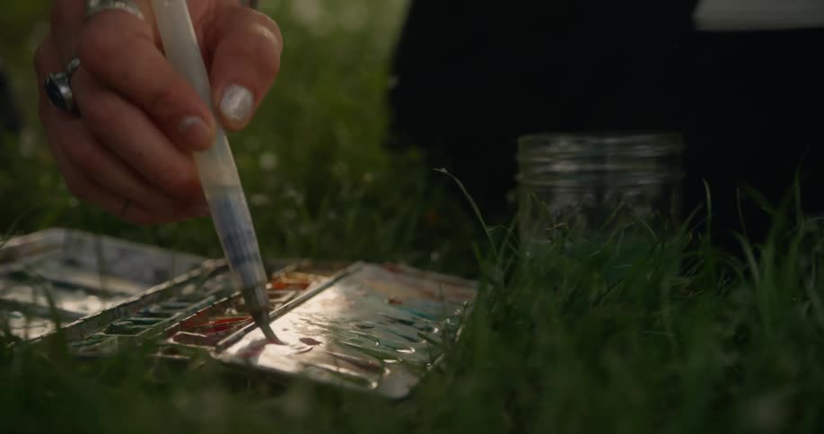 Hand with modern paintbrush tinting a paint palette which is on top of the grass outdoors