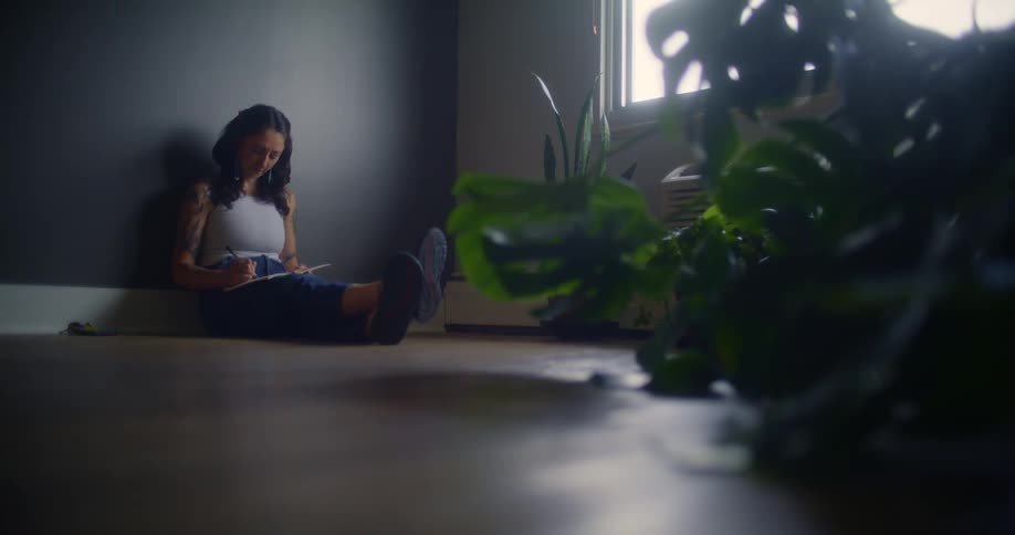 A woman in a white sleeveless shirt and blue jeans sits on the living room floor leaning against a dark wall while writing in a notebook with soft natural light filtering through a nearby window and houseplants adding a touch of greenery