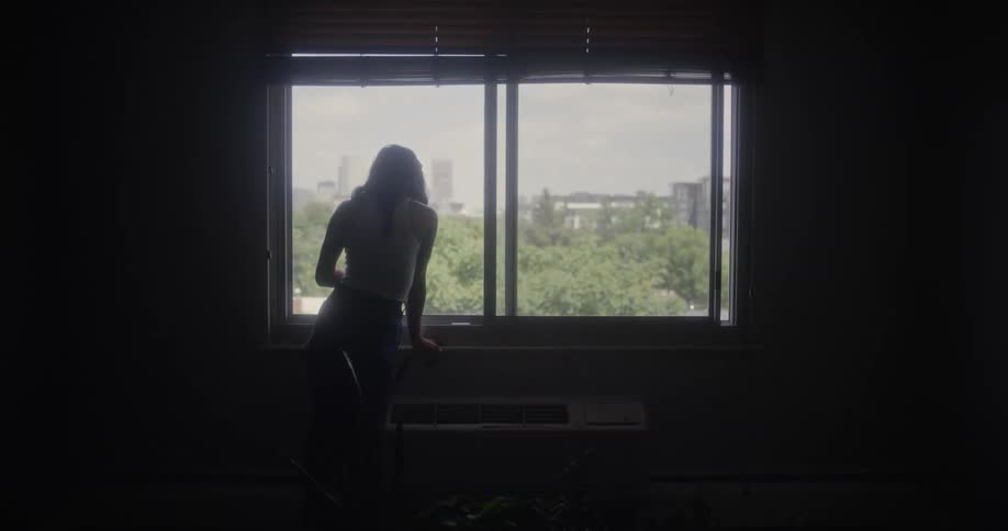 A woman stands in a dimly lit room leaning to look out a window at the sunny city