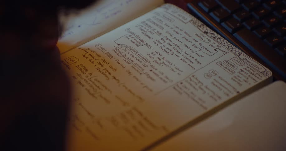 A notebook filled with handwritten notes and diagrams lies open next to a computer keyboard in a dimly lit setting